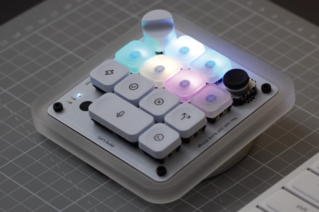 Um teclado modular branco com teclas iluminadas em tons de azul, roxo e branco, sobre uma base quadriculada cinza. Algumas teclas têm ícones como microfone, sigma e setas. Há um joystick preto no canto superior direito e texto Lets build e You can just build things na base