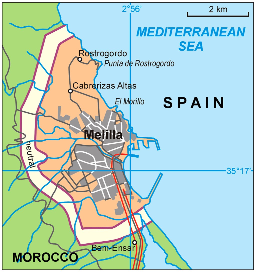 Mapa de Melilla, cidade autônoma espanhola na costa do Mar Mediterrâneo, cercada por Marrocos. A área urbana cinza destaca-se na península laranja, com estradas e o porto. A fronteira com Marrocos é marcada por uma linha roxa, e uma área neutra clara separa os países. Coordenadas geográficas e escala de 2 km estão visíveis