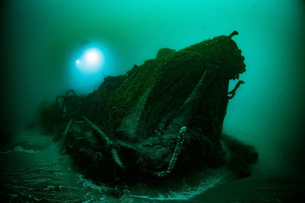 Um naufrágio escuro e coberto de algas no fundo do mar, iluminado por um feixe de luz azul-clara vindo da esquerda, em águas verde-azuladas turvas