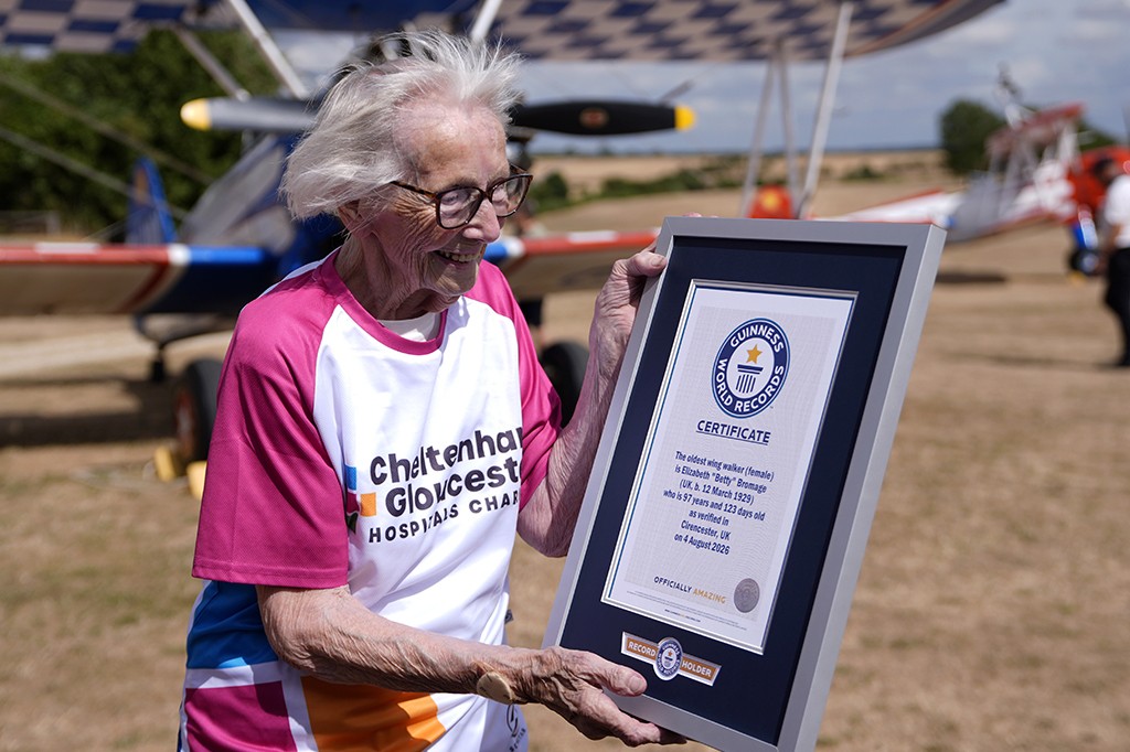 Mulher idosa de cabelos brancos e óculos, sorrindo e segurando um certificado do Guinness World Records, com um avião ao fundo. Ela veste uma camiseta branca e rosa com a inscrição Cheltenham & Gloucester Hospitals Charity. O certificado reconhece Elizabeth Betty Bromage como a wing walker (andadora de asa) mais velha, aos 97 anos e 123 dias, em 4 de agosto de 2026