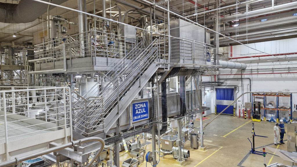 Interior de uma fábrica moderna com equipamentos industriais de aço inoxidável, tubulações e plataformas elevadas. Uma escada metálica central leva a um nível superior. Um letreiro azul na estrutura indica ZONA AZUL (BLUE ZONE). No chão, dois trabalhadores com capacetes e uniformes brancos e azuis observam o ambiente. O piso é amarelo com faixas de segurança, e ao fundo há uma porta azul e prateleiras com materiais