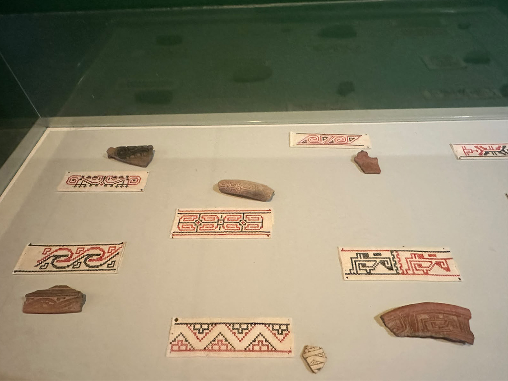Exposição de artefatos indígenas com peças de cerâmica marrom e tecidos bordados em branco, vermelho e preto, exibindo padrões geométricos e espirais em uma vitrine de museu