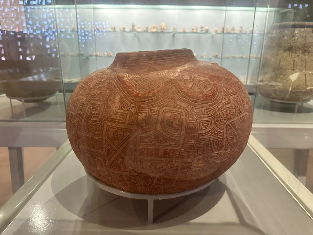 Vaso de cerâmica marrom-avermelhada com padrões geométricos intrincados gravados em sua superfície, exibido em um museu