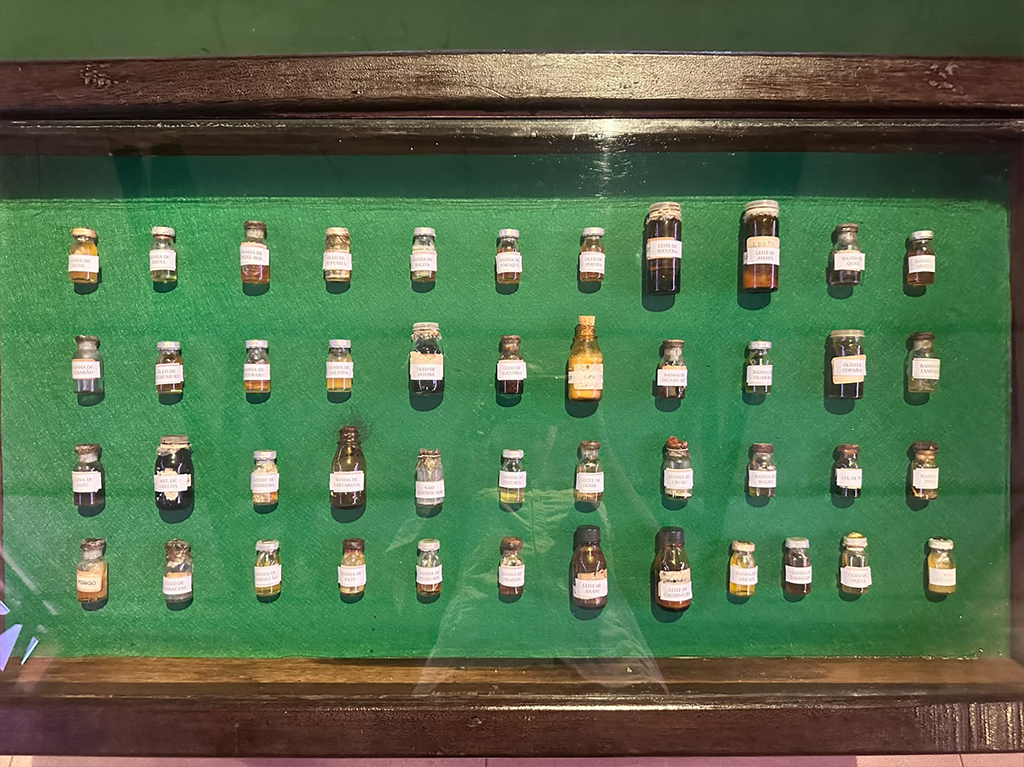 Exposição de quarenta e cinco pequenos frascos de vidro com líquidos e substâncias variadas, organizados em fileiras sobre um fundo verde dentro de uma caixa de madeira escura com tampa de vidro