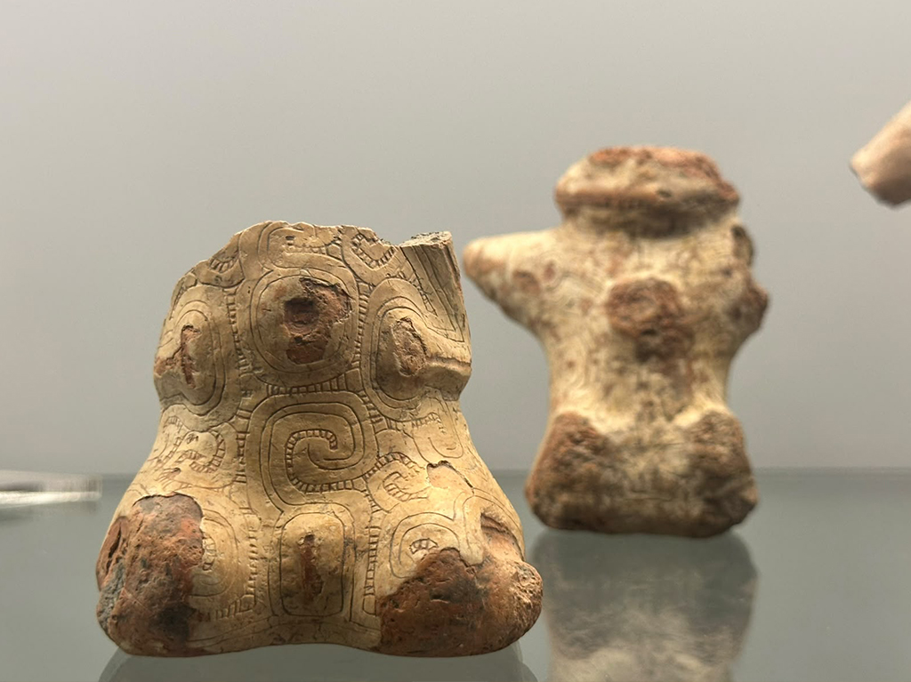 Duas estatuetas de cerâmica antiga em tons de bege e marrom, com a da frente detalhada com padrões espirais e geométricos, sobre uma superfície reflexiva