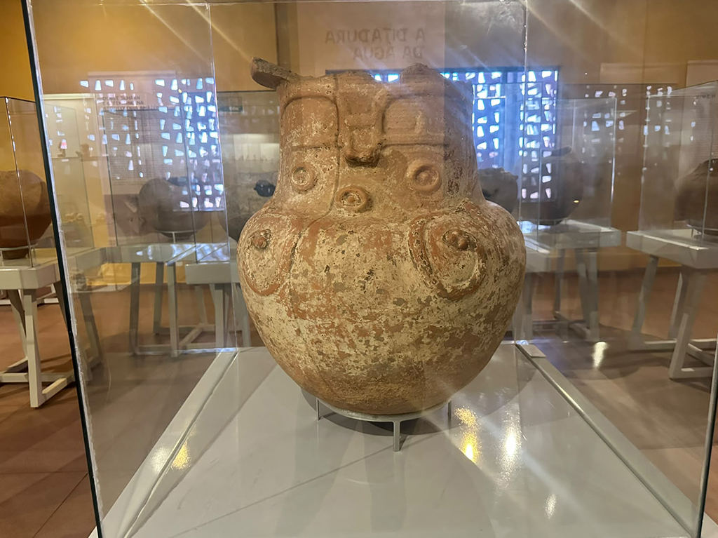 Vaso de cerâmica antigo, marrom-claro e desgastado, com rosto estilizado e detalhes em espiral, exposto em vitrine de vidro em um museu