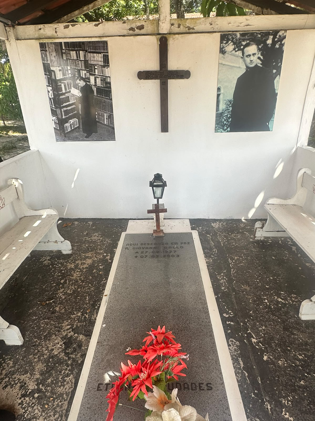 Túmulo do Padre Giovanni Gallo, com lápide cinza e flores vermelhas. Na parede branca, duas fotos em preto e branco do padre e uma cruz de madeira. Bancos brancos ladeiam o túmulo
