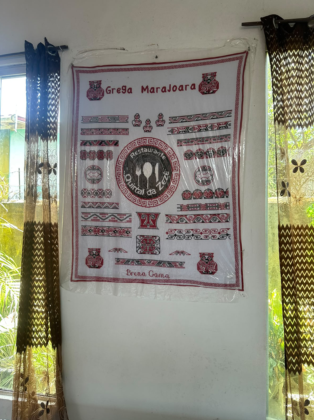 Pano branco com bordados marajoaras em vermelho e preto, exibindo o texto Grega Marajoara e abaixo, Brena Gama. O pano está fixado em uma parede branca, entre duas janelas com cortinas estampadas