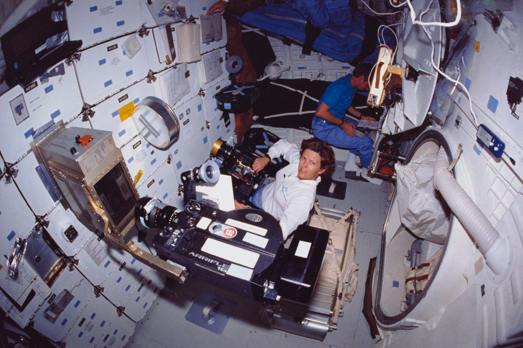 Dois astronautas em um módulo espacial branco. Um homem de camisa branca e calça azul sorri para a câmera enquanto opera uma grande câmera de vídeo preta. Outro homem de camisa azul e calça azul está sentado ao fundo, concentrado em um laptop. O ambiente é repleto de equipamentos e painéis de controle fixados nas paredes.