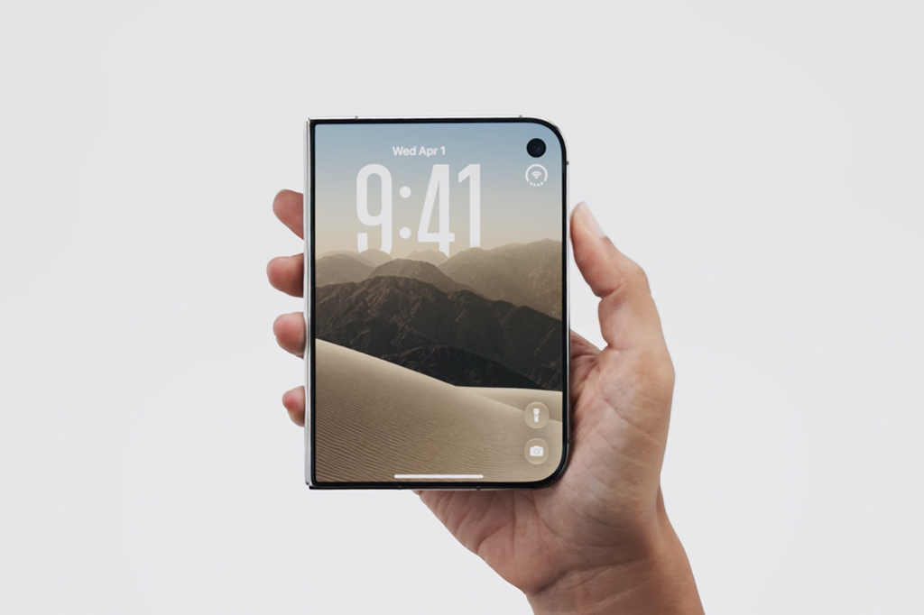 Mão segurando um smartphone dobrável com tela ligada exibindo a hora 9:41, data Wed Apr 1 e um papel de parede de dunas e montanhas desérticas