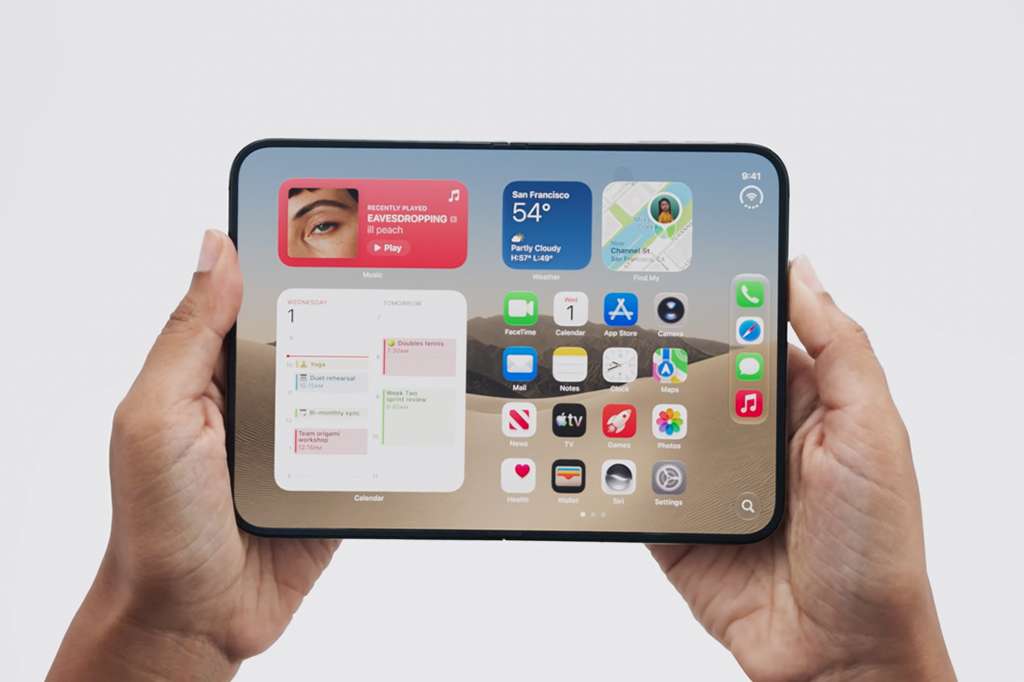 Duas mãos seguram um tablet horizontalmente, exibindo a tela inicial com widgets de música, clima, calendário e vários ícones de aplicativos sobre um fundo de dunas de areia