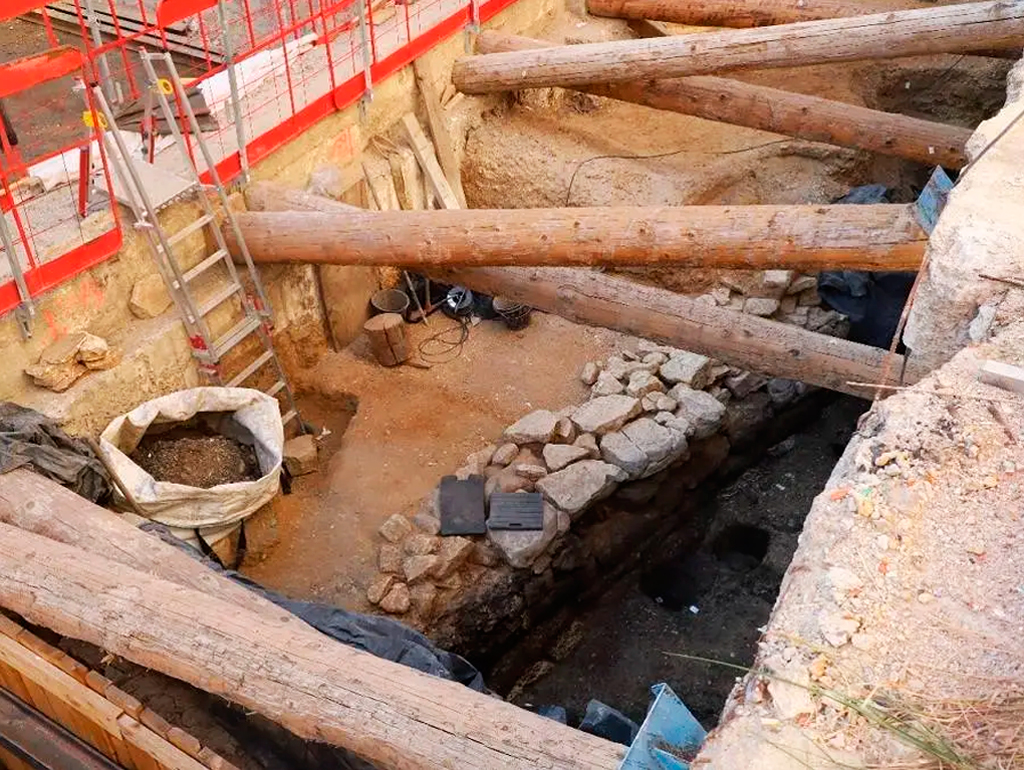 Canteiro de obras com escavação profunda, escorada por troncos de madeira e cercada por grades de segurança vermelhas. No fundo, um saco de ráfia cheio de terra e pedras, com uma escada de alumínio apoiada na parede de terra.