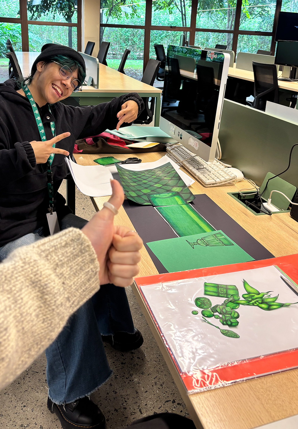 Uma pessoa sorridente com gorro preto e óculos redondos aponta para desenhos verdes sobre uma mesa de madeira, enquanto outra pessoa faz um sinal de positivo com o polegar. Há um monitor de computador e janelas com árvores ao fundo