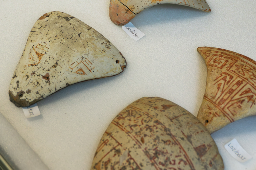 Tanga de cerâmica antiga com padrões geométricos em tons de ocre e marrom sobre fundo claro, exibindo marcas de identificação em etiquetas brancas, dispostos sobre uma superfície branca