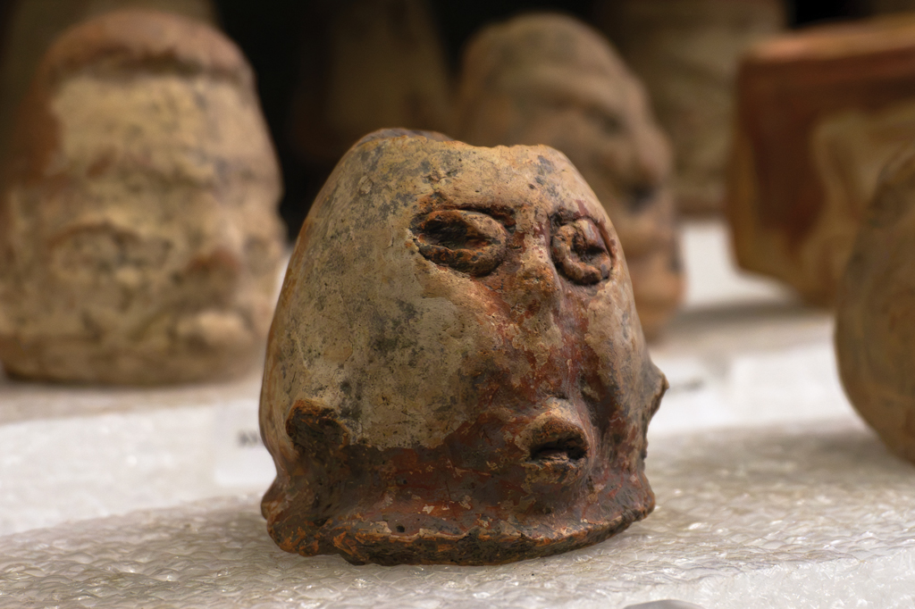 Chocalho de argila antiga em formato de cabeça humana, com olhos e boca marcados, em tons terrosos de bege, marrom e vermelho, sobre plástico bolha, com outras peças desfocadas ao fundo