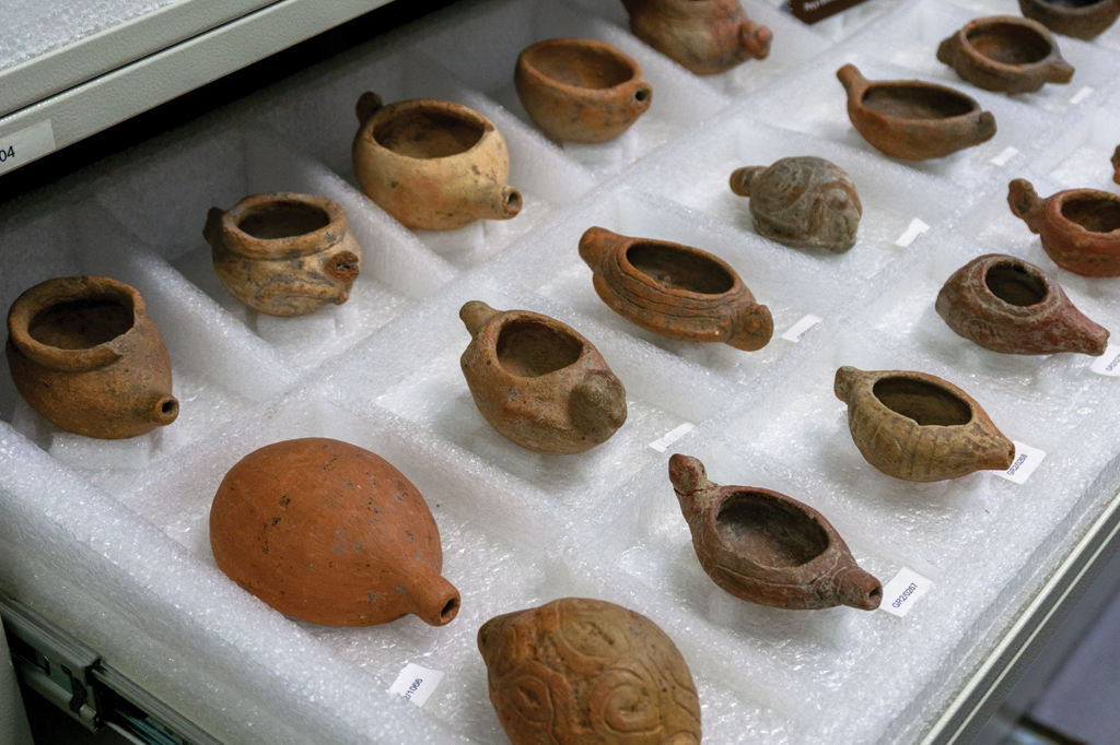 Várias rapezeiras antigas e vasos de cerâmica em tons terrosos, dispostas em fileiras sobre plástico bolha branco dentro de uma gaveta de museu. Algumas peças têm etiquetas de identificação brancas