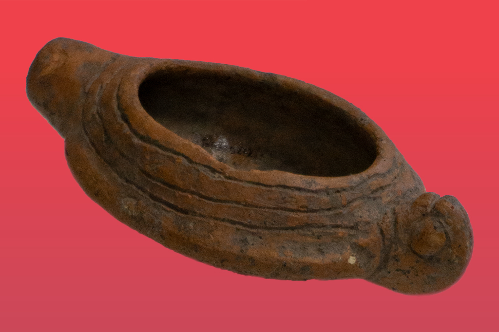 Antiga rapezeira de barro marrom-avermelhado, com formato oval alongado e bico saliente, apresentando sulcos horizontais decorativos na lateral e uma cavidade central para o óleo, sobre fundo vermelho