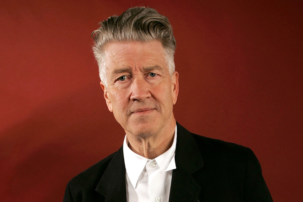 Cineasta David Lynch, h omem branco de meia-idade, com cabelo grisalho penteado para trás, vestindo camisa branca e blazer preto, olhando diretamente para frente com expressão séria, contra um fundo vermelho escuro