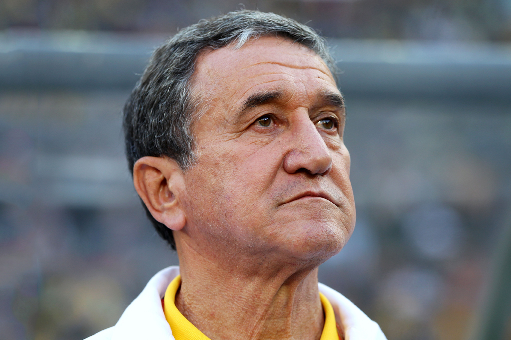 Técnico de futebol Carlos Alberto Parreira, homem de meia-idade, cabelos grisalhos e pele bronzeada, com expressão séria e olhar para cima, vestindo camisa polo amarela e casaco branco, em um ambiente externo.