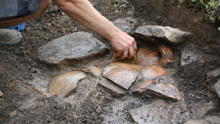Um arqueólogo com calça jeans e sapato sujo, ajoelhado, limpa cuidadosamente com a mão um artefato de cerâmica marrom-avermelhada, parcialmente submerso em água barrenta, em uma escavação de terra escura com pedras ao redor