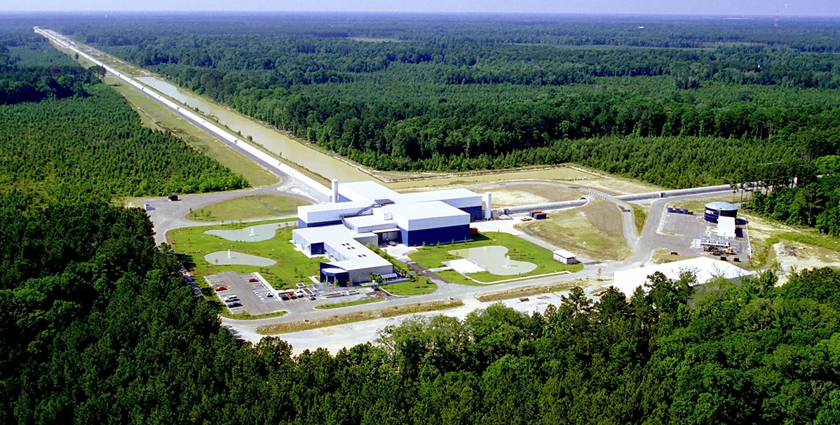 Vista aérea do Observatório de Ondas Gravitacionais por Interferômetro Laser (LIGO) em Livingston, Louisiana, com edifícios brancos e azuis no centro, cercados por vastas áreas de floresta verde. Dois braços longos e retos, com cerca de 4 km cada, estendem-se da estrutura central, formando um L e desaparecendo na distância. Há pequenos lagos e estacionamentos com carros visíveis perto dos edifícios. O céu é azul claro